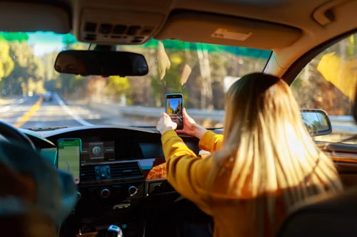 Driver holding a smartphone while driving on a sunny road, with dashboard and rearview mirror visible