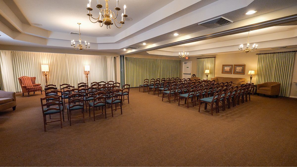 Rows of chairs face a curtained stage in a carpeted room, with chandeliers and sconces.