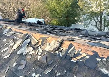 A roofer removing old asphalt shingles from a roof. The roof is partially stripped, revealing the underlayment.