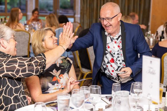A man in a suit and patterned shirt high-fives a woman at a table during a brightly lit indoor event.