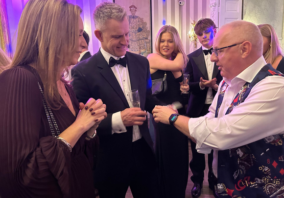 A man in a patterned vest performs a card trick for an audience at an indoor evening event.
