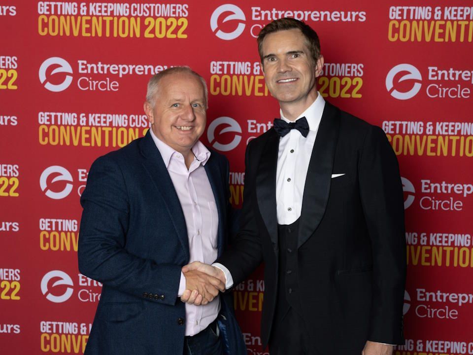 Peter Barker and Jimmy Carr are shaking hands in front of a sign that says Entrepreneurs Convention