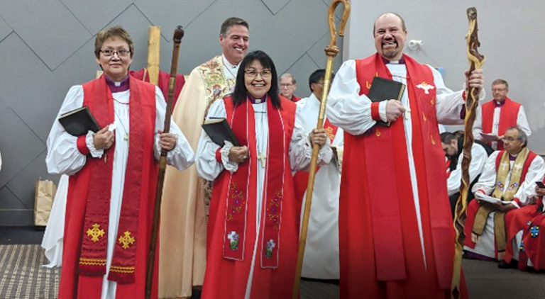 Clergy in red robes stand together, holding Bibles and staffs, smiling. Interior setting, church.
