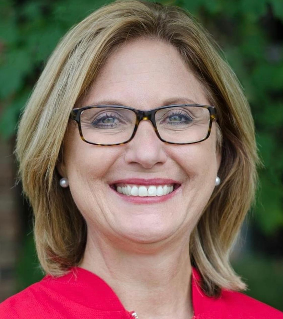 Woman with glasses smiling, wearing a red top, with a blurred outdoor background.