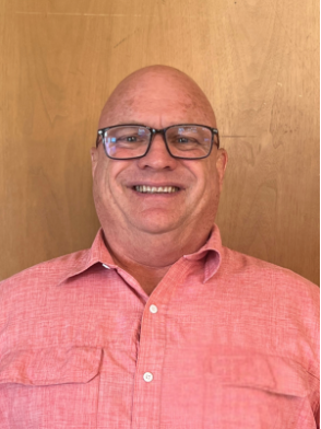 Bald man with glasses smiles, wearing a pink button-down shirt, against a wood panel background.