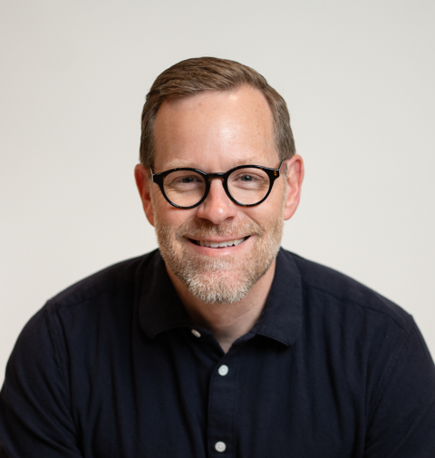 Man with glasses smiles at the camera. He has short brown hair and a navy blue shirt.