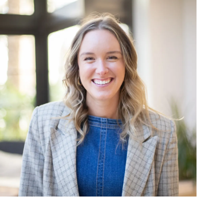 Woman with blonde hair, wearing a denim top and plaid blazer, smiling in a bright setting.