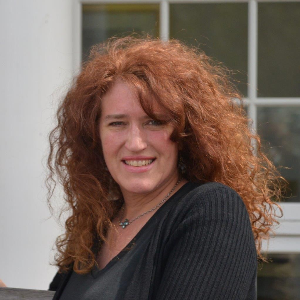 Woman with curly red hair smiles, wearing black, in front of a building.