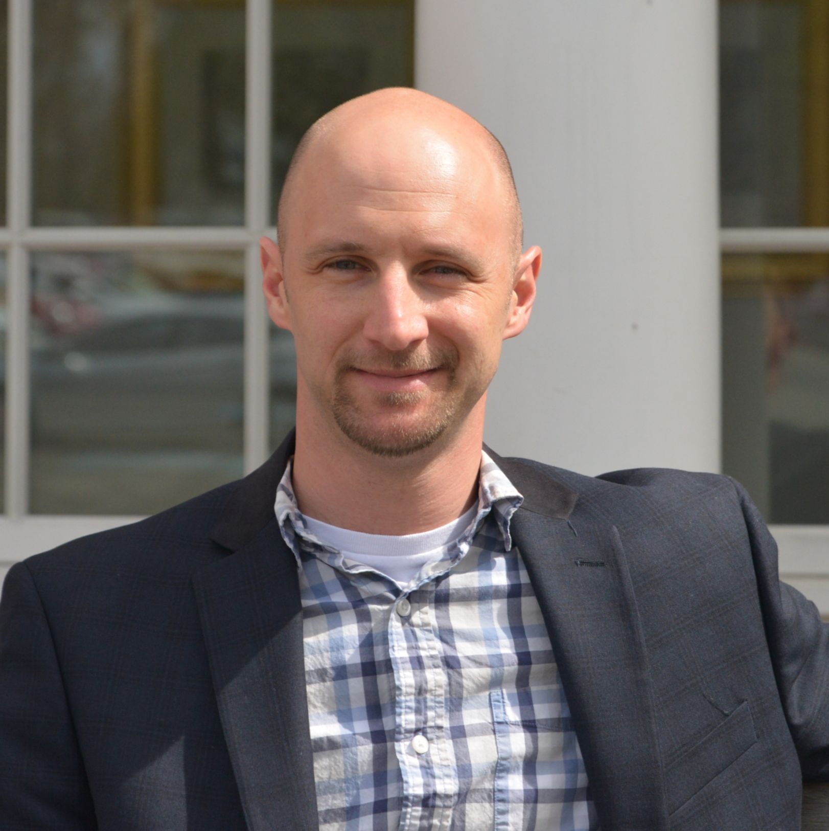 Bald man wearing a blazer and plaid shirt, smiling, outdoors with a building in the background.