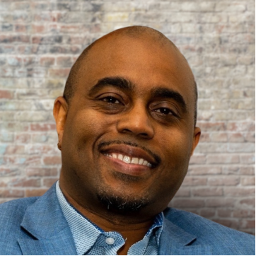 Smiling man in a blue suit with a brick background.