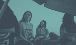 Group of women outdoors, smiling and looking up, under a canopy with a cloudy sky.