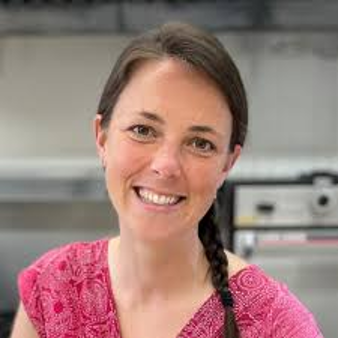 Woman in a pink top smiles, brown braid, in a kitchen.