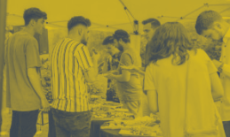 People browsing an outdoor market, some examining items at a table under a canopy. Yellow tint.