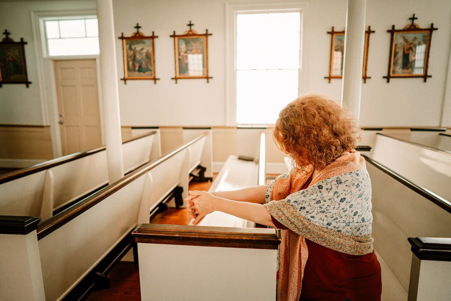 Woman in a church, hands clasped in prayer. Warm light, paintings on the wall, light-colored pews.