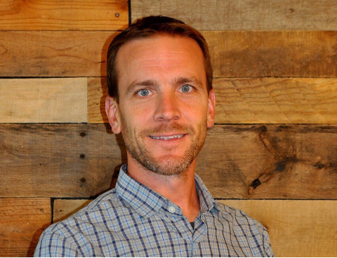 Man with short brown hair, blue eyes, and a light beard, smiles in front of a wooden wall.