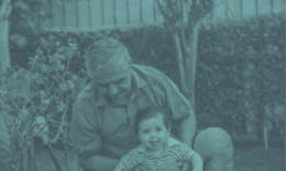 A grandfather smiles while holding a baby outdoors.
