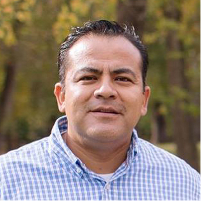 Man with dark hair and a blue plaid shirt smiles outside.