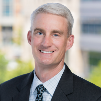 Middle-aged man with fair skin, graying hair, smiling in a suit. Background shows a blurred cityscape.