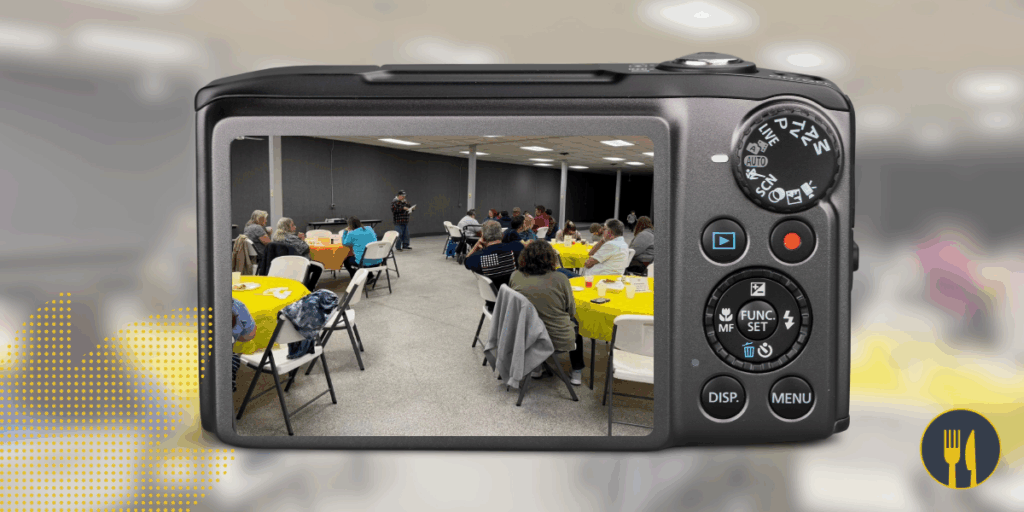 A camera displaying a room with people at tables, possibly at a meeting or event.