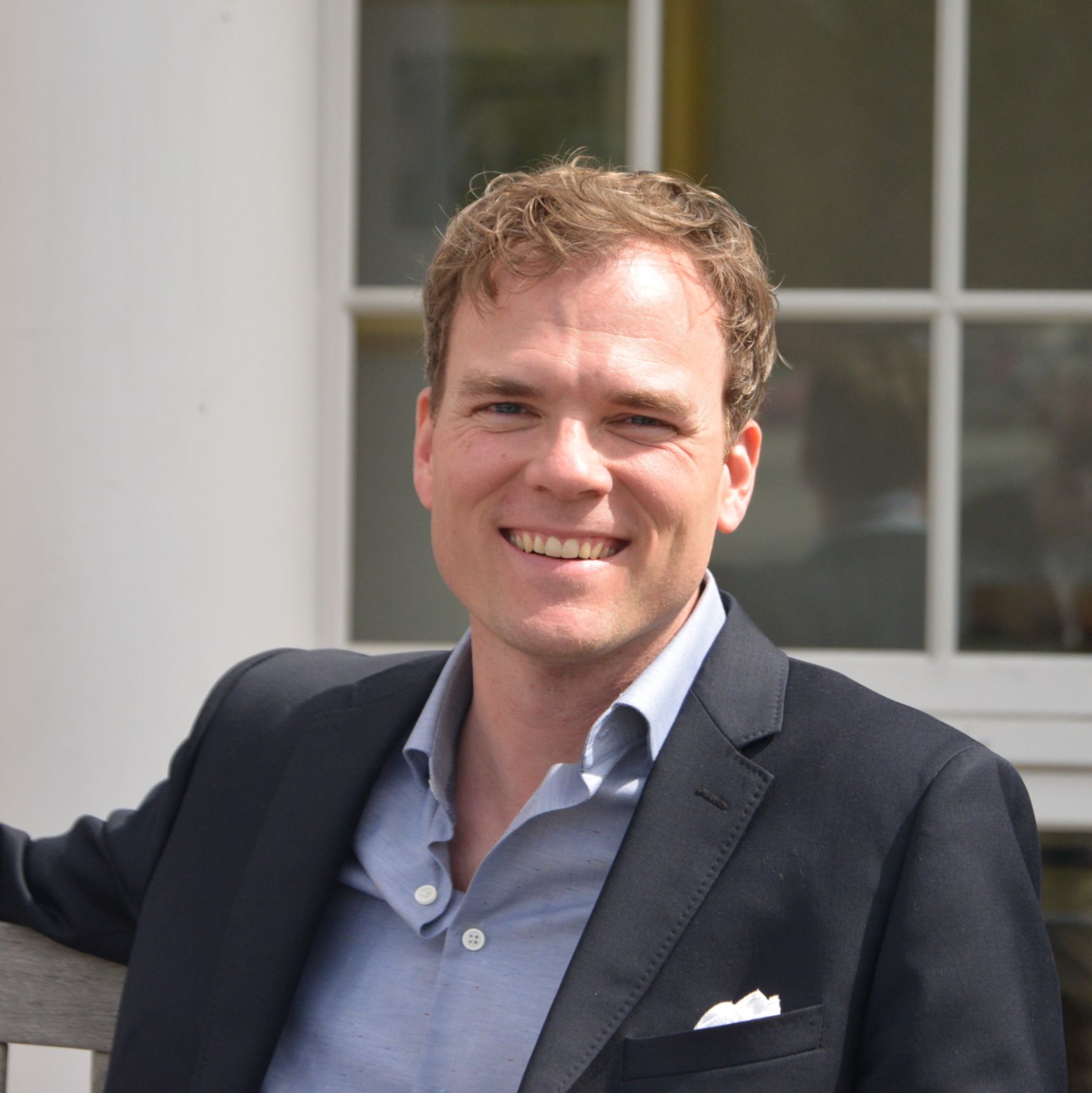 Man in dark blazer smiles, seated outdoors near a building with white pillars and windows.