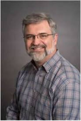 Smiling man with gray hair and beard, wearing glasses and a plaid shirt.