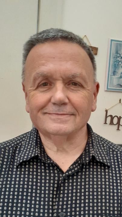 Middle-aged man with graying hair smiles, wearing a black patterned shirt, in a home setting.