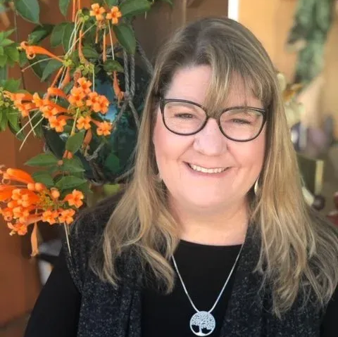 Smiling woman with glasses, wearing a silver pendant, with orange flowers in the background.