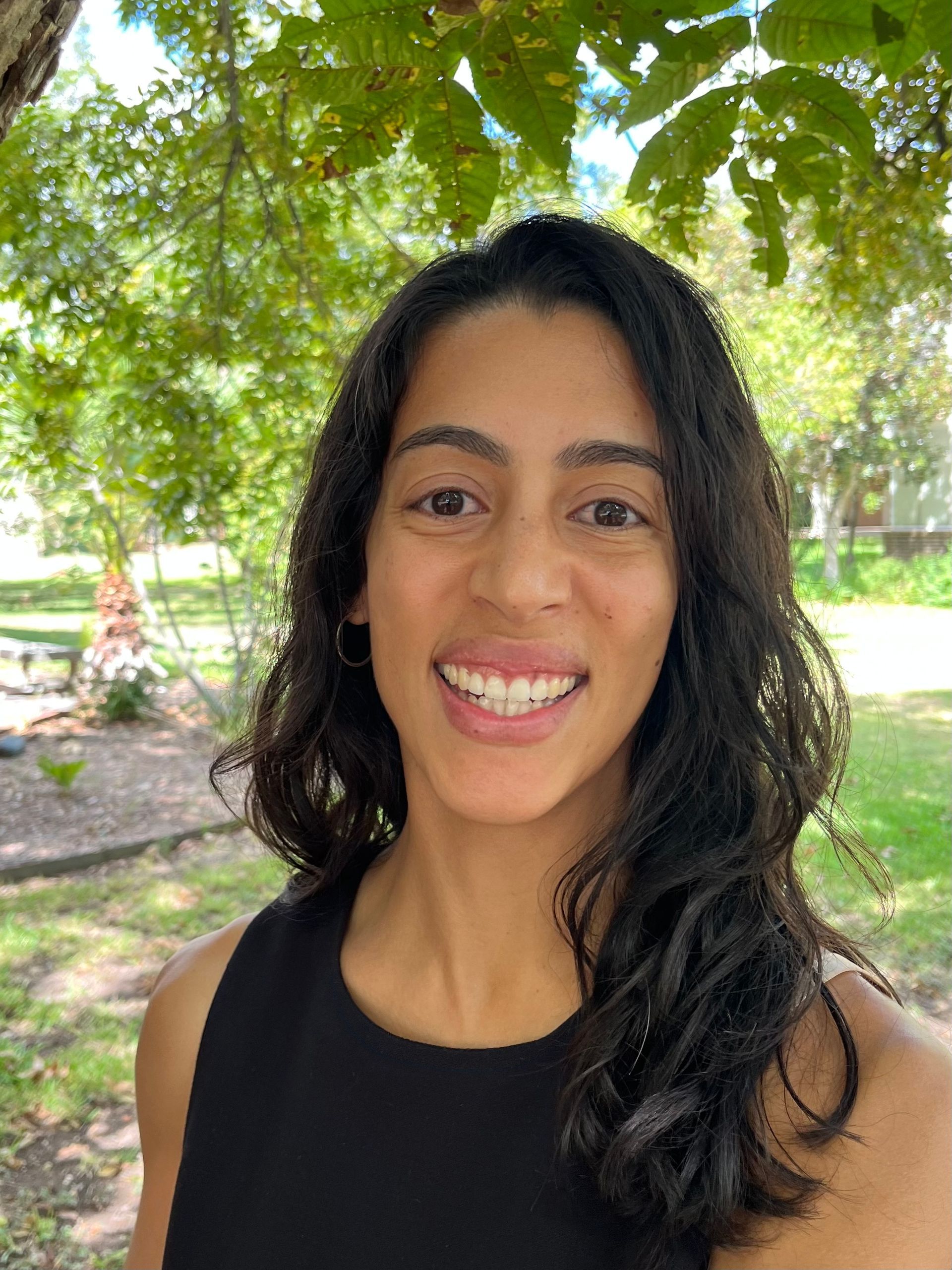 Woman with dark hair smiling, outdoors near a tree, wearing a black top.