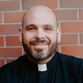A smiling, bald man with a beard wearing a clerical collar, in front of a brick wall.