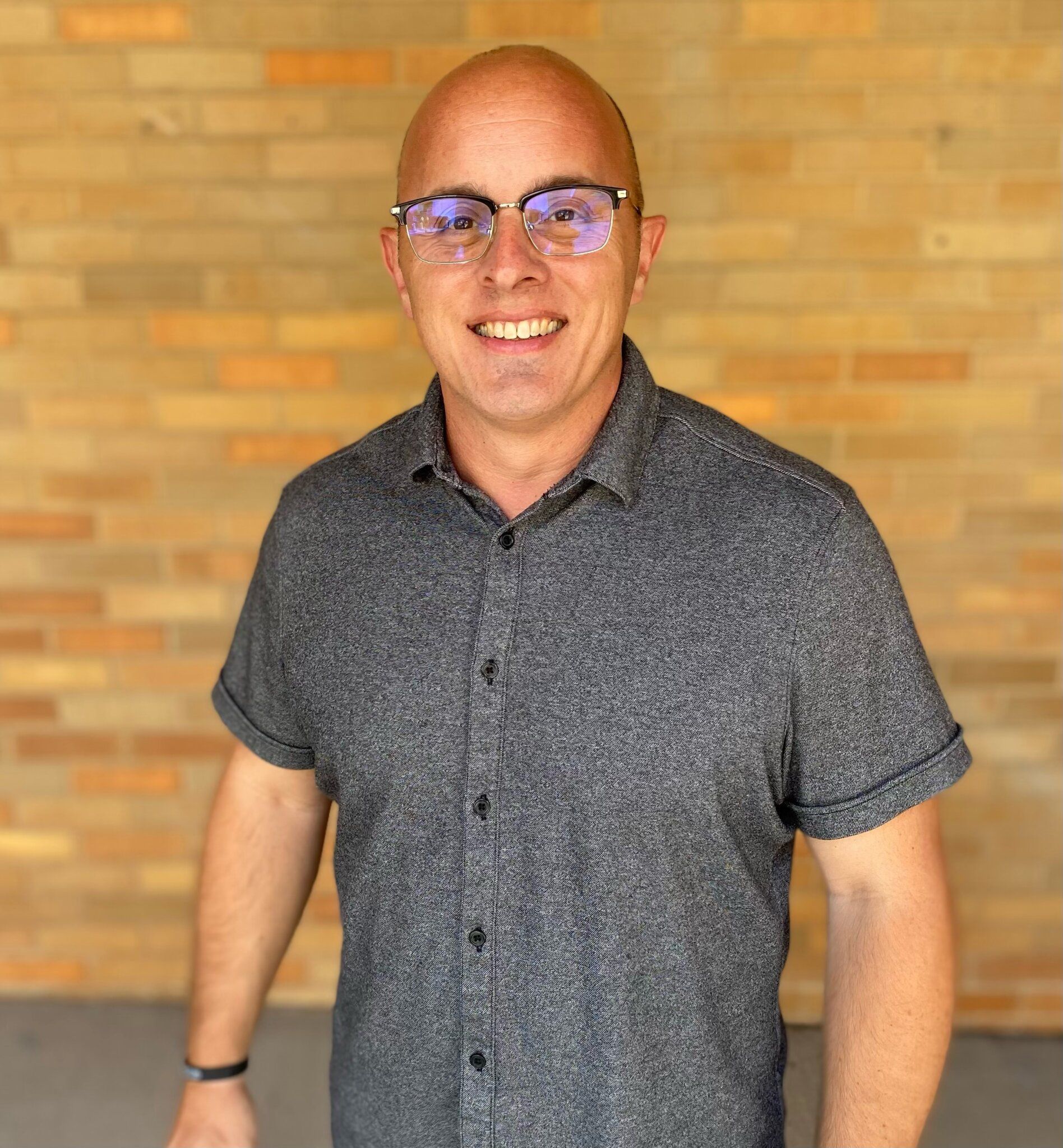 Bald man with glasses smiles in front of a brick wall, wearing a gray button-down shirt.