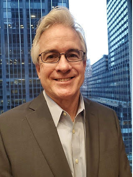 Man with glasses in a suit smiles in front of a city skyline.