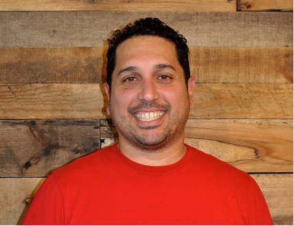 Man in red shirt smiles in front of a wood-paneled wall.