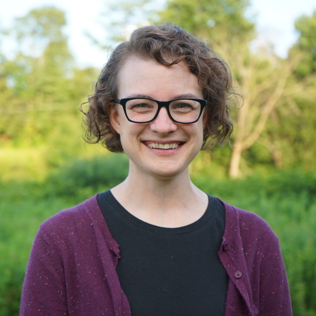 Woman with glasses smiles outdoors in a purple sweater, black shirt, and curly hair. Green trees and grass in background.