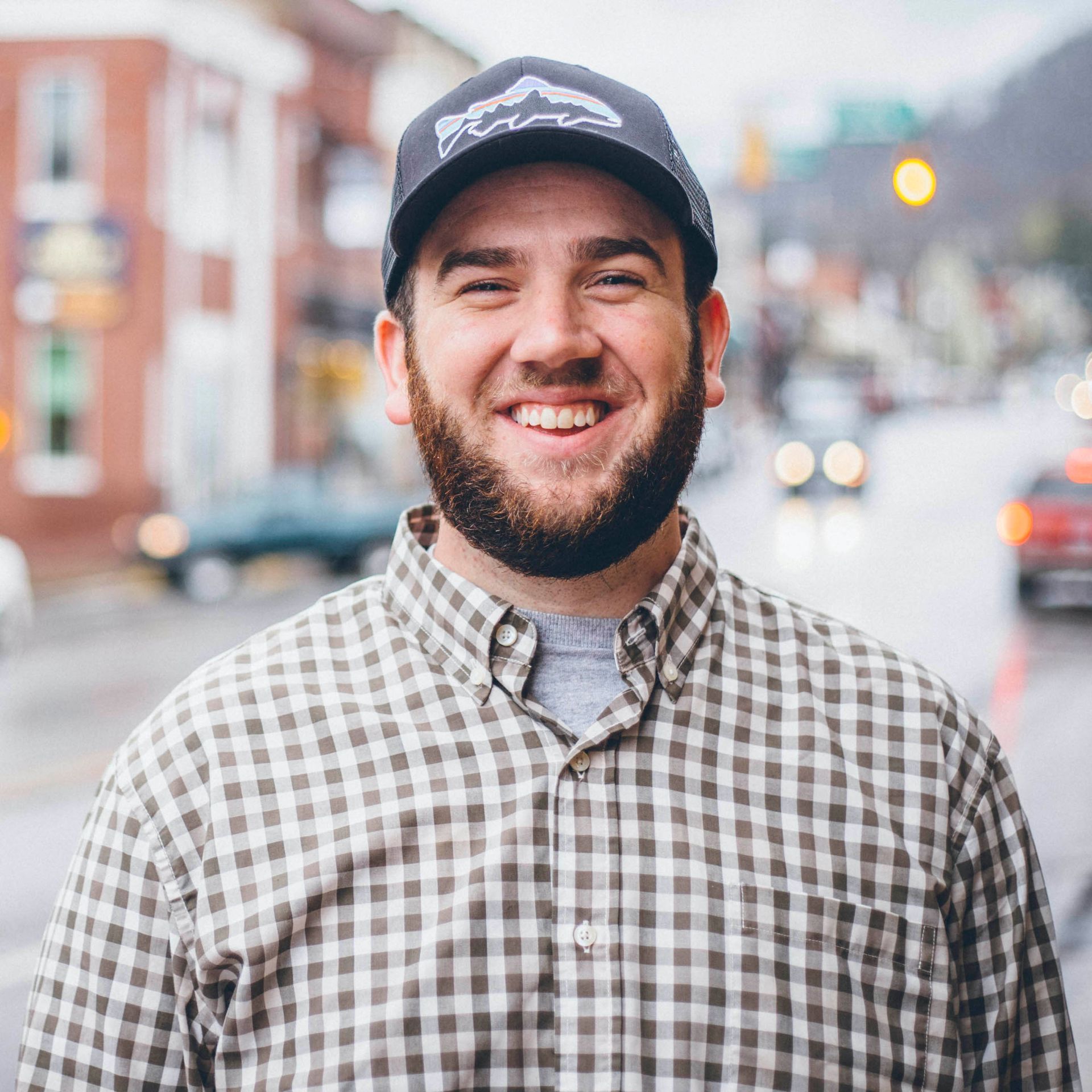 Man in glasses and hat smiles, wearing a striped shirt, in front of a city.