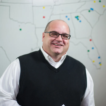 Man in glasses and black sweater vest smiles, posing in front of a map of the US with colorful pins.