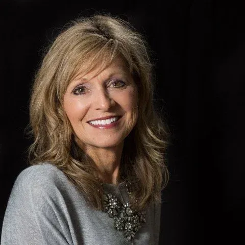 Woman with blonde hair, smiling, wearing a gray sweater and necklace, against a black background.