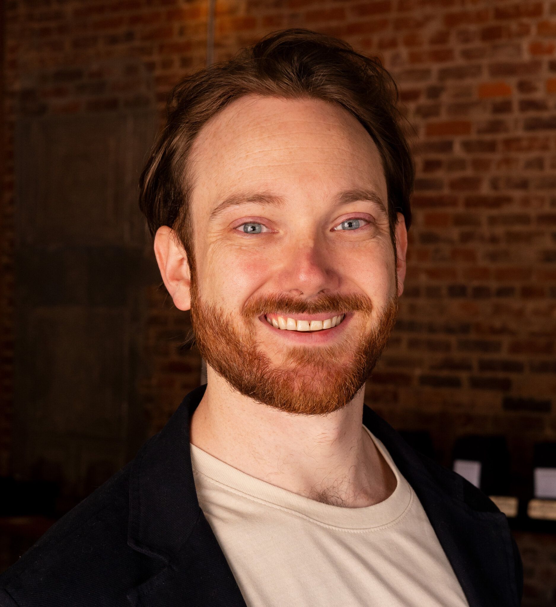 Man with red hair and beard smiles at the camera against a brick wall background.