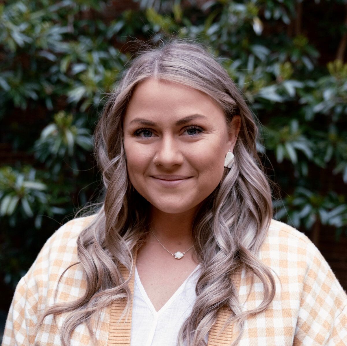 Woman with blonde hair smiles, wearing a yellow checkered jacket and white shirt, outdoors near green foliage.