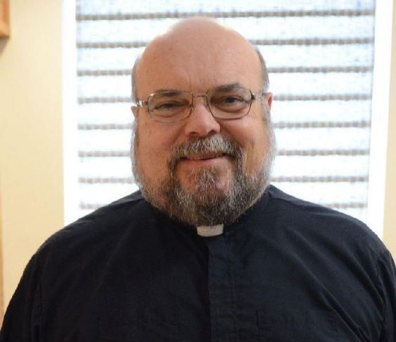 A man with glasses, a beard, and clerical collar smiles at the camera.
