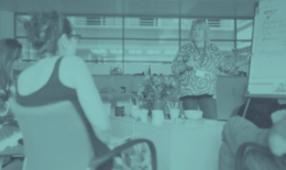 Woman presents at meeting; other attendees watch in office setting.