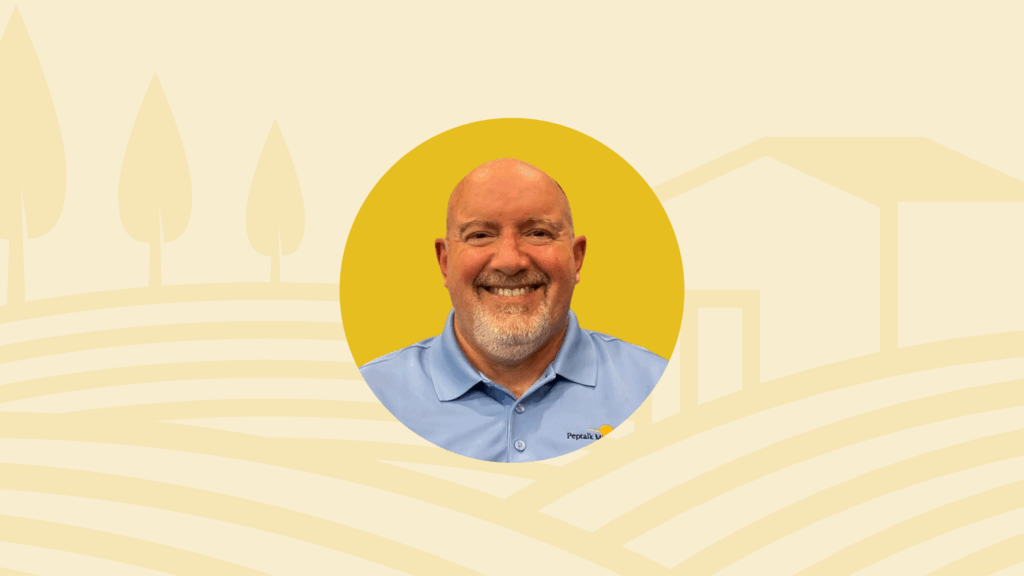 Smiling bald man with a beard, in a blue shirt, inside a yellow circle, against a light yellow farm background.