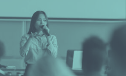 Woman giving a presentation with a microphone in front of an audience in a classroom.