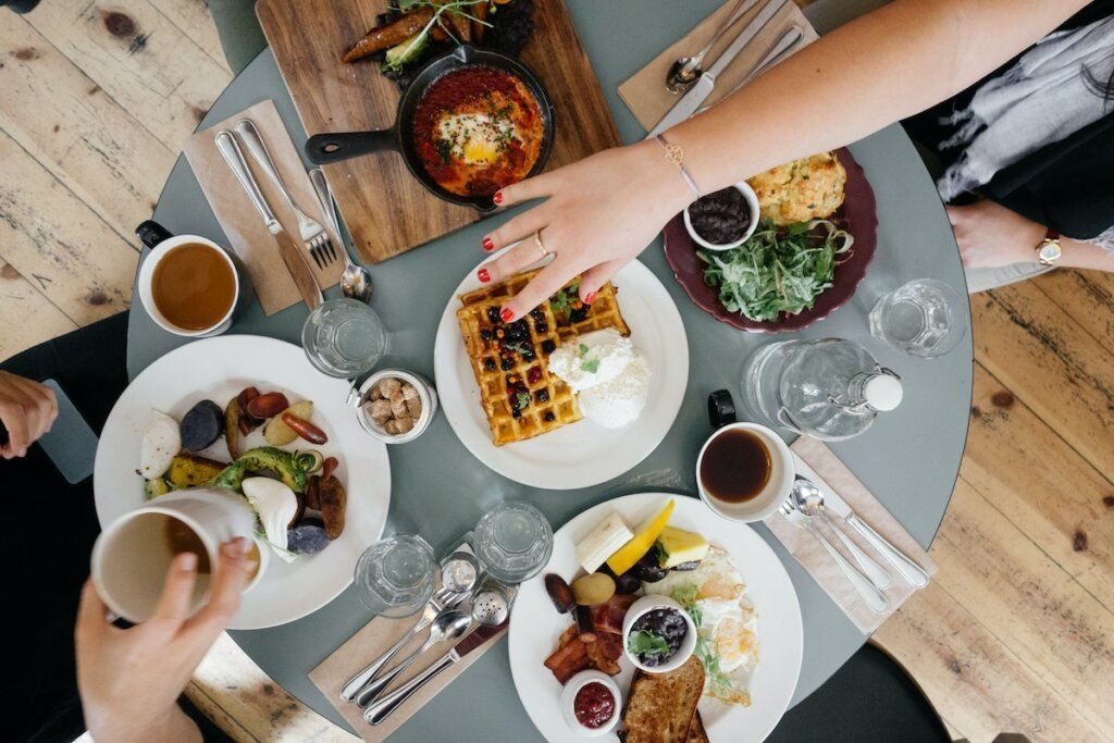 Overhead view of a brunch table with plates of food, coffee cups, and people's hands reaching for the food.