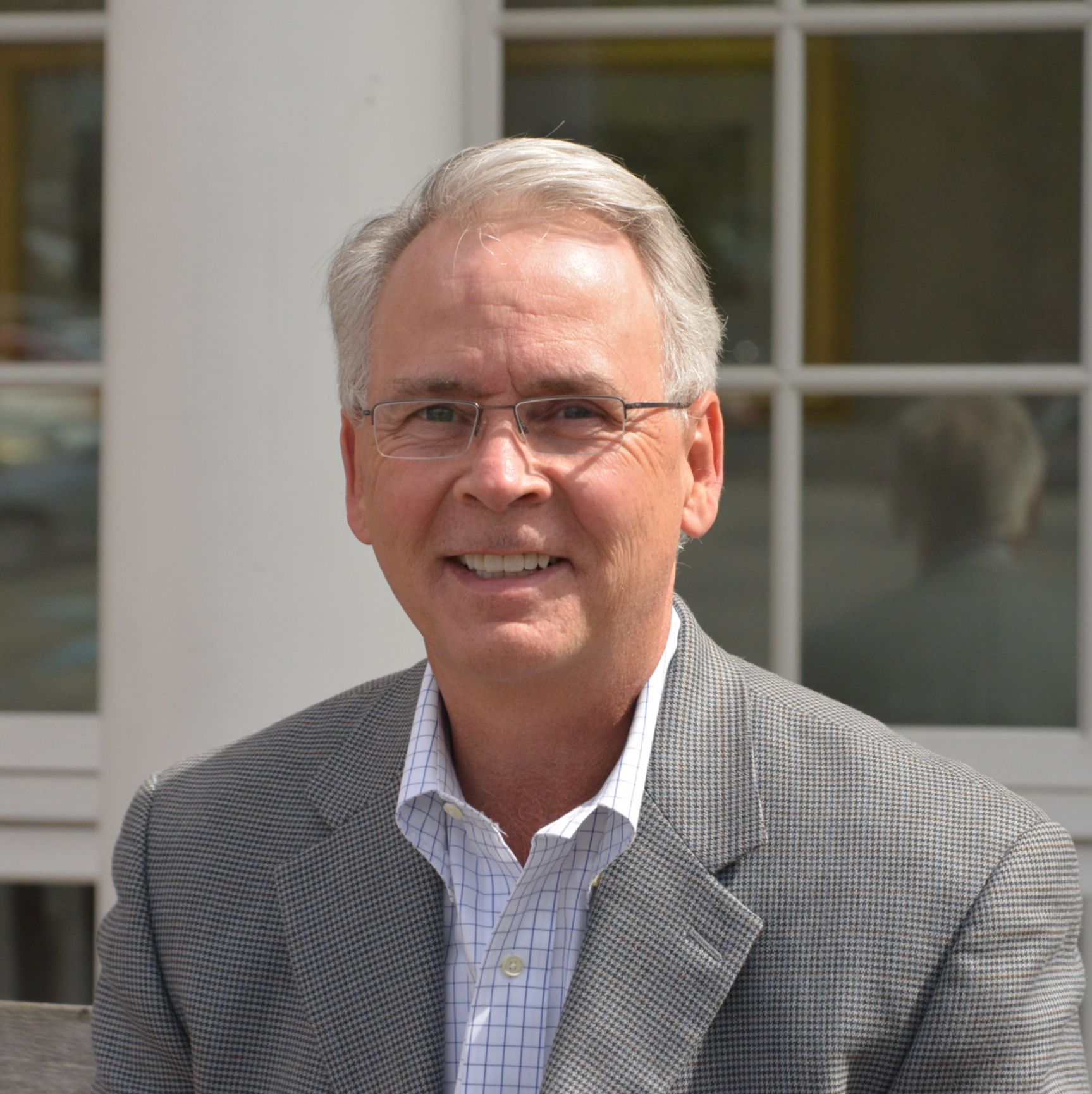 Man in glasses smiles, wearing a gray blazer and light blue shirt, in front of a building.