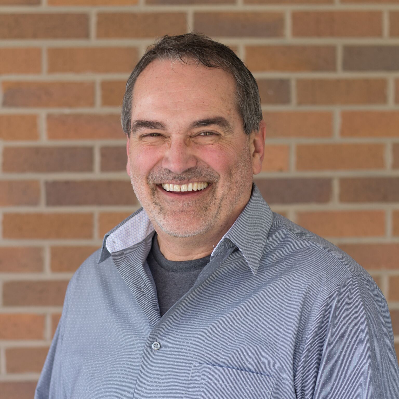 Smiling man with graying hair and beard in front of a brick wall.