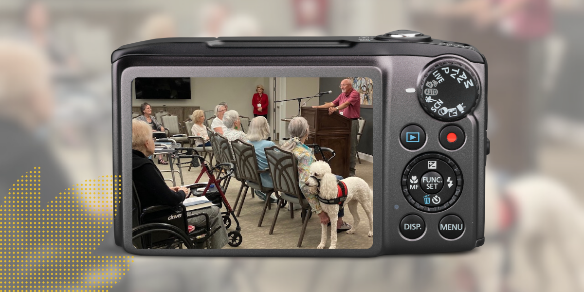 Camera displaying a room of elderly people, some using walkers, listening to a speaker; a white dog is present.