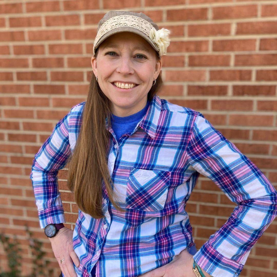 Woman with long brown hair in plaid shirt and visor smiles in front of a brick wall.