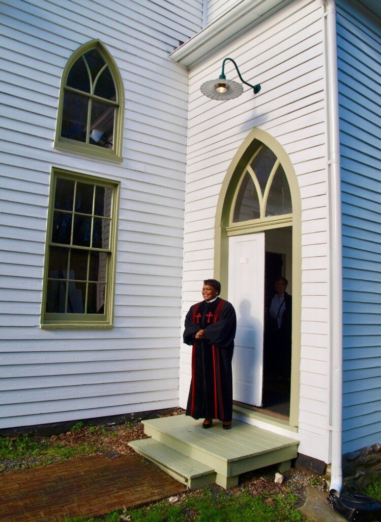 Man in robes stands at church doorway. White building, green trim, arched windows.