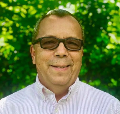 Man with glasses smiles, wearing a pink striped shirt, against a green leafy background.