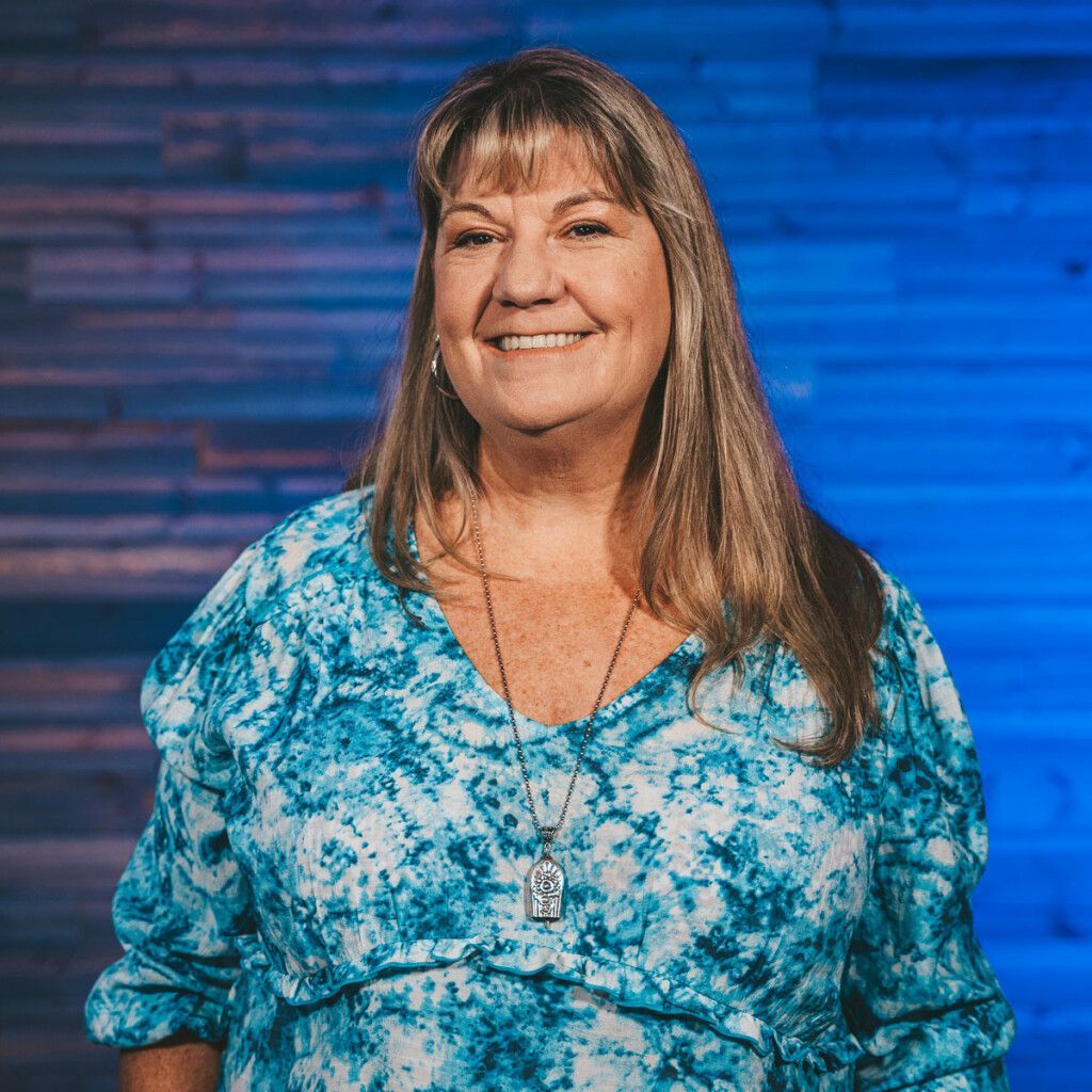 Woman with long blonde hair, smiling in a blue tie-dye top. Blue background.